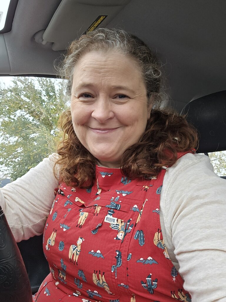 Smiling lady with greying red hair wearing red alpaca print overalls and a white shirt